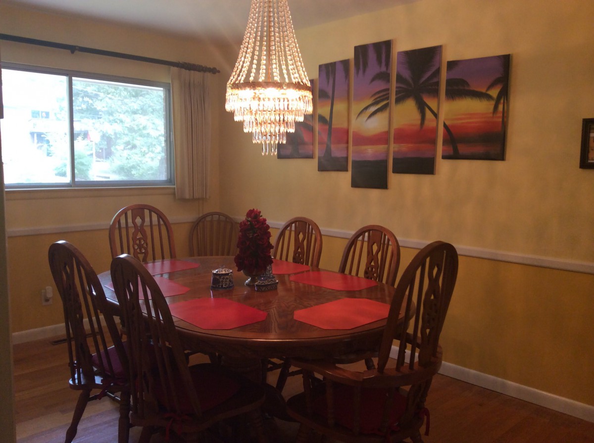 A cozy dining room featuring a wooden dining table with red placemats, surrounded by chairs and illuminated by a chandelier. The wall is adorned with a colorful piece of artwork depicting palm trees.