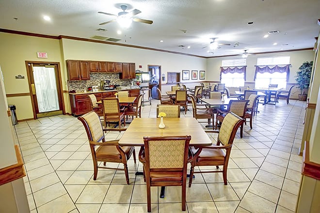 A spacious dining area with neatly arranged tables and chairs, featuring a tiled floor and kitchen facilities in the background.