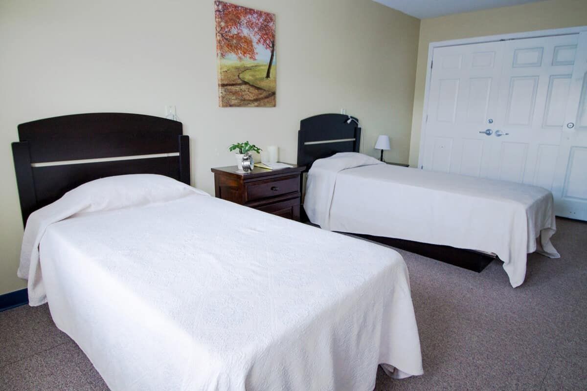 A cozy bedroom featuring two single beds with white bedspreads, a nightstand with a lamp and decorative items, and a painting on the wall.