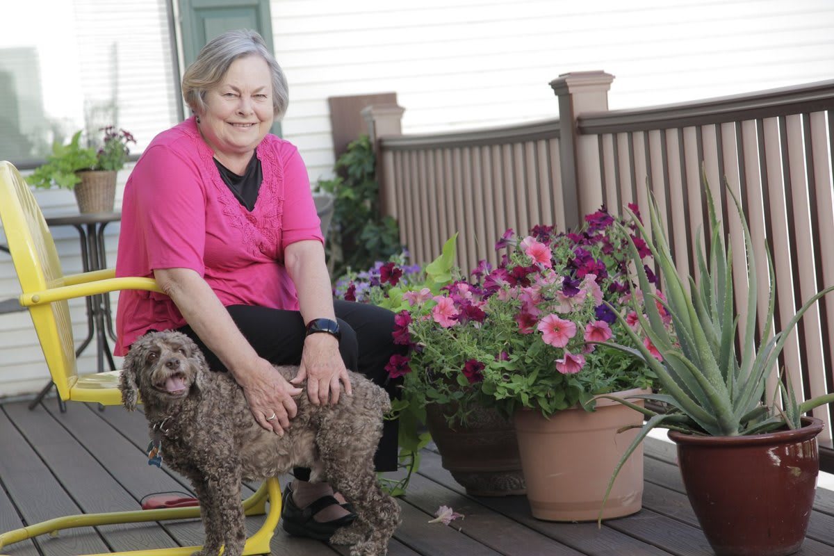 A woman sitting on a porch with a dog beside her, surrounded by potted flowers.