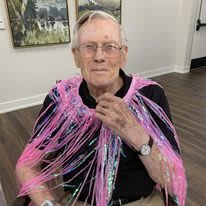 Elderly man wearing a pink decorative fringe indoors.
