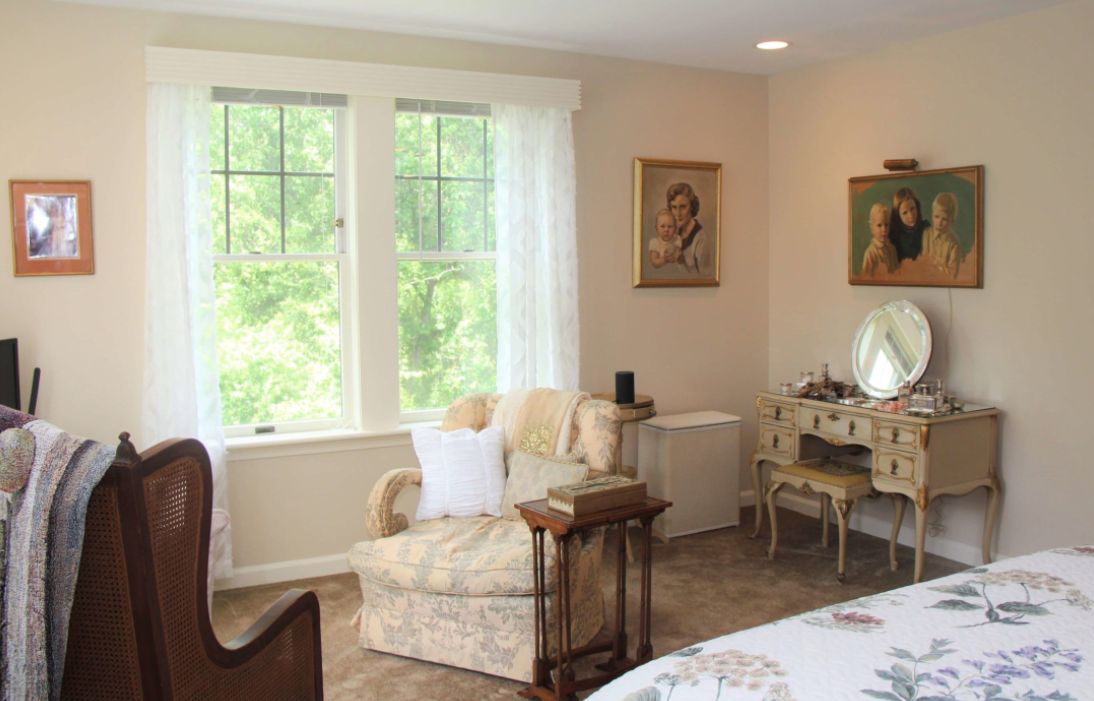 A cozy living room with vintage decor, featuring an armchair, artwork on the walls, and a bright window view.