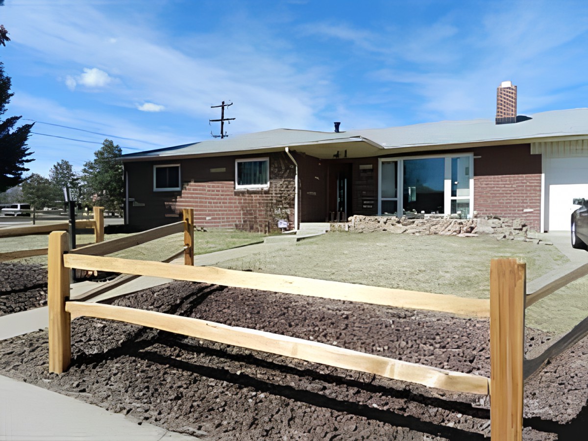 A single-story brick house with a front lawn and wooden fence, set against a clear blue sky.