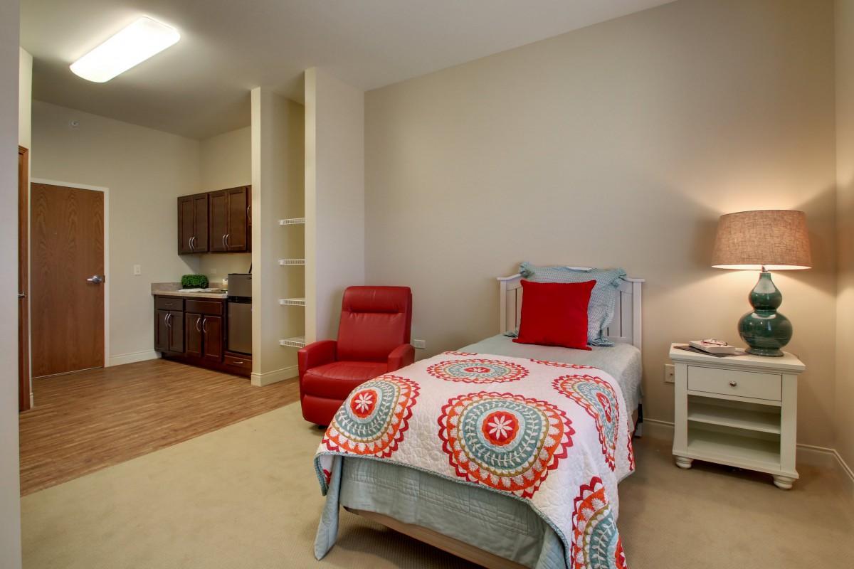 A cozy bedroom with a single bed and armchair next to a small kitchenette.