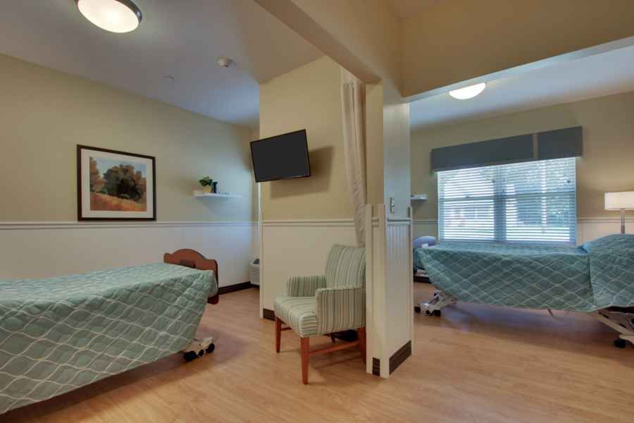 A hospital room with patient beds separated by curtains, featuring a television and chairs.