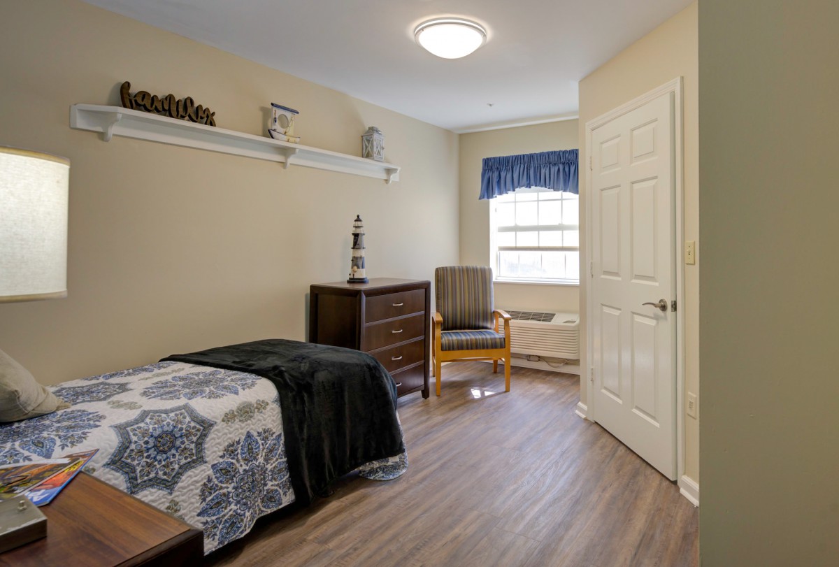 A neatly arranged bedroom with a single bed, dresser, and chair by the window.