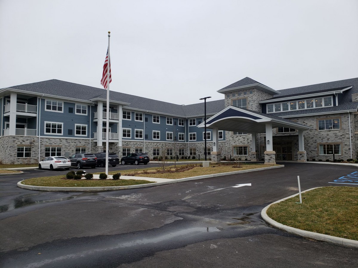 A large building with an American flag in front, surrounded by a parking lot and driveway on a cloudy day.