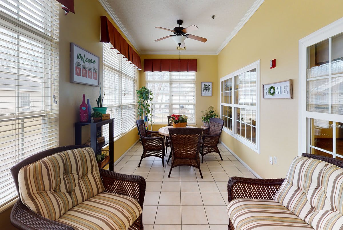 A cozy indoor seating area with wicker furniture, large windows, and a ceiling fan.