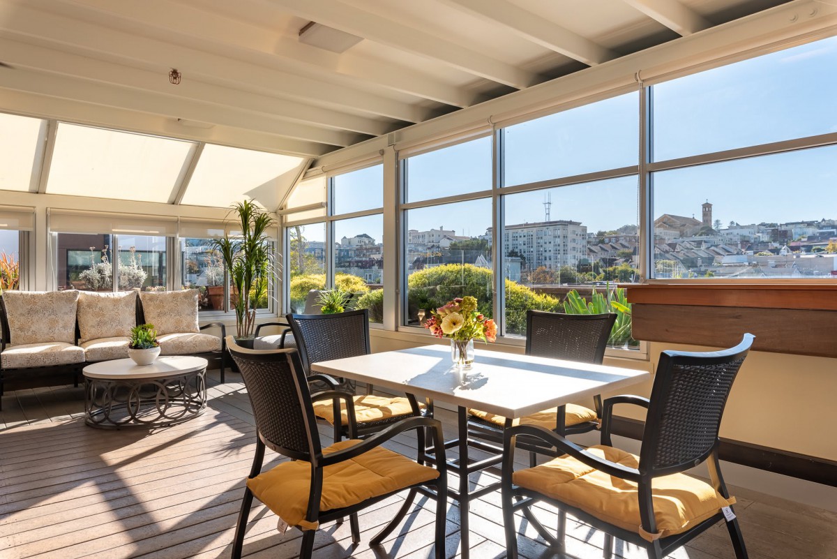 A sunny patio with a table and chairs, surrounded by plants, offering a view of the cityscape.