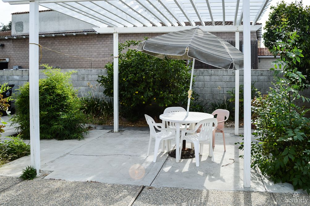 A backyard patio featuring white plastic chairs and a table under a pergola, with an umbrella for shade.