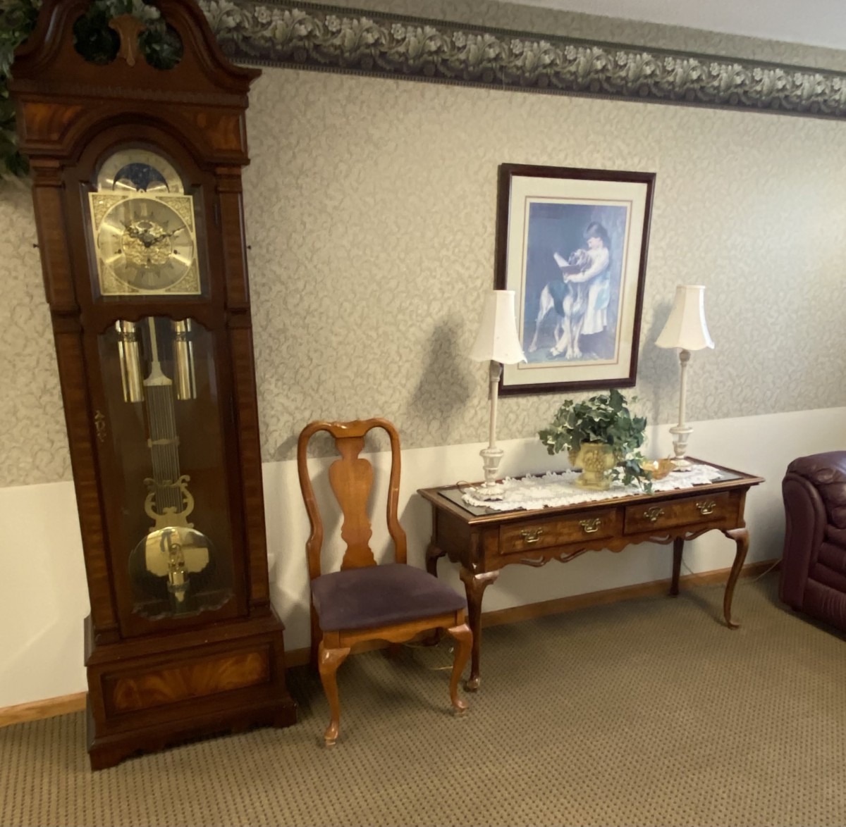 A cozy room corner featuring a grandfather clock, a wooden chair, and a table topped with decorative items beneath wall art.