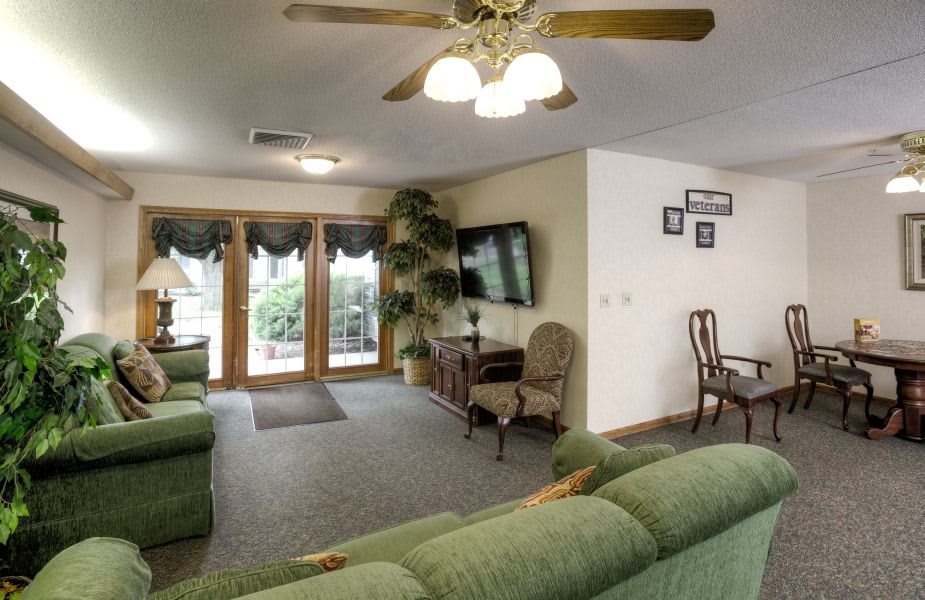 A cozy living room with green sofas, an armchair, a wall-mounted TV, and a ceiling fan.