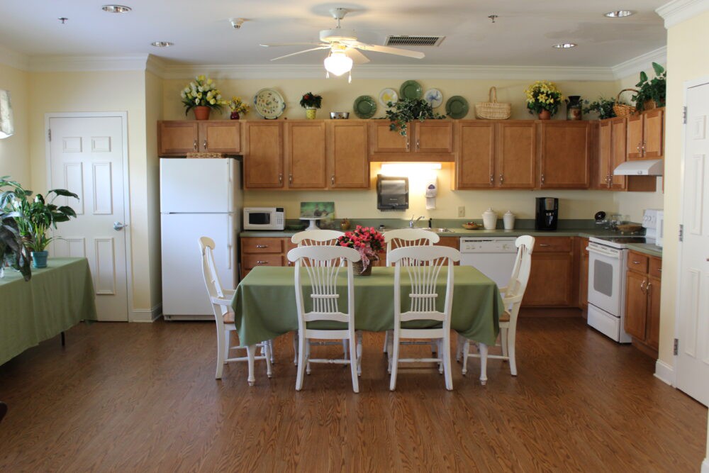 A cozy kitchen and dining area featuring wooden cabinets, a refrigerator, a microwave, and a table set with chairs. The space is decorated with plants and flowers.