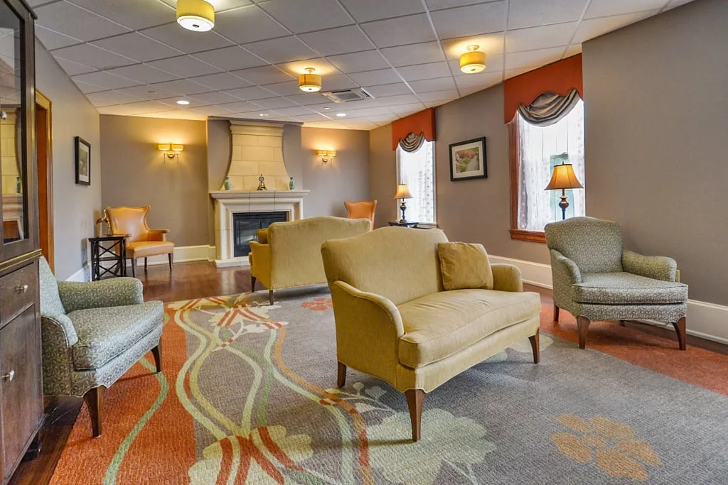 A cozy lounge area with armchairs and sofas arranged around a fireplace, featuring large windows and elegant decor.