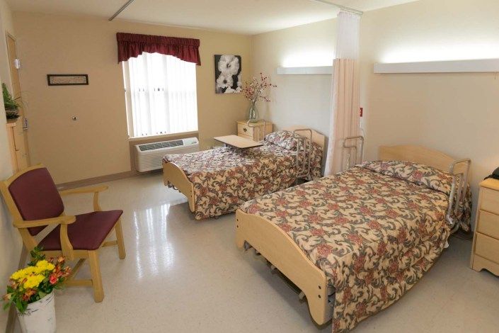 A shared room with two beds, decorated in a floral pattern, featuring a window with curtains and basic furniture.