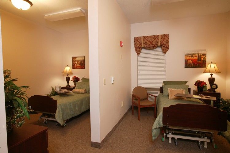 A shared room with two hospital beds separated by a partial wall, featuring bedside tables and chairs.