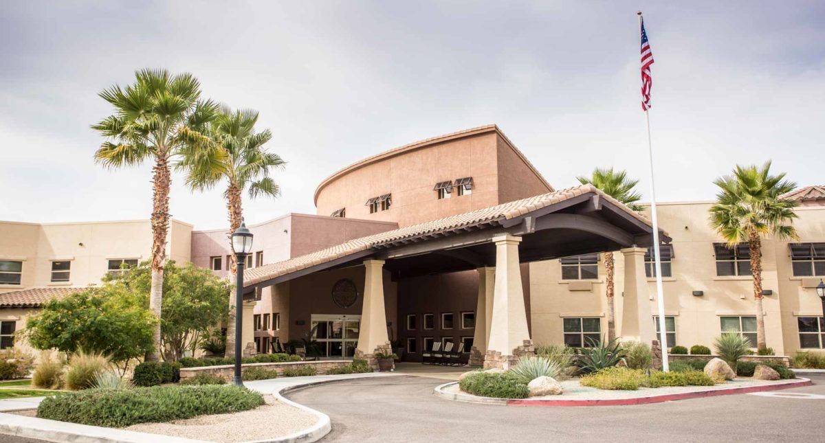 The image shows the entrance of a modern building surrounded by palm trees under a cloudy sky.