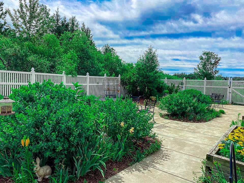 A serene garden with lush greenery, a pathway, and a wooden fence under a partly cloudy sky.