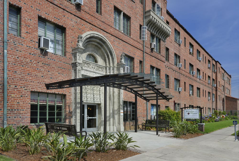 Entrance of a brick apartment building with a metal canopy and landscaped surroundings.