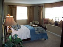 A cozy bedroom featuring a bed with blue and white bedding, two windows with curtains, and a sitting area by the window.