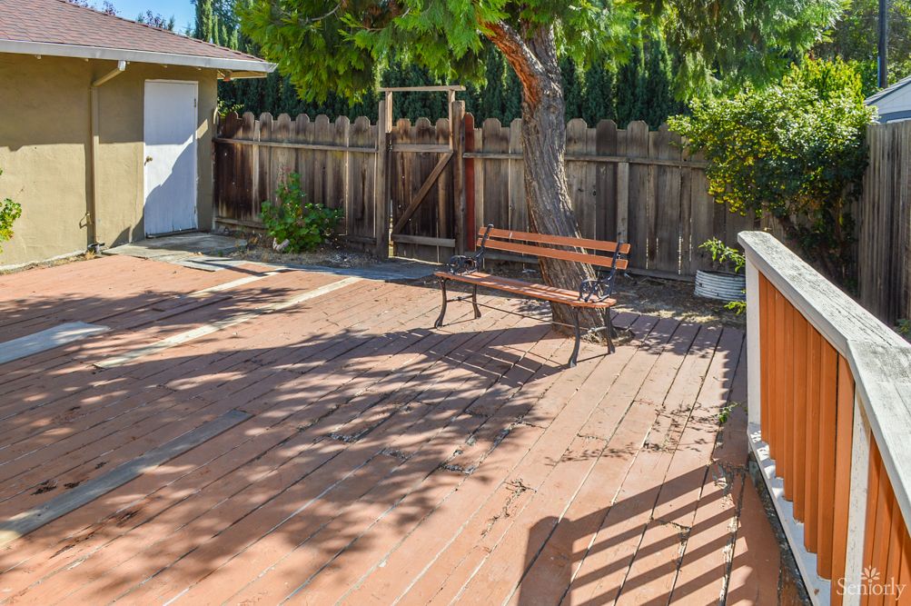 A sunny backyard with a wooden deck, featuring a bench and tree surrounded by a fence.