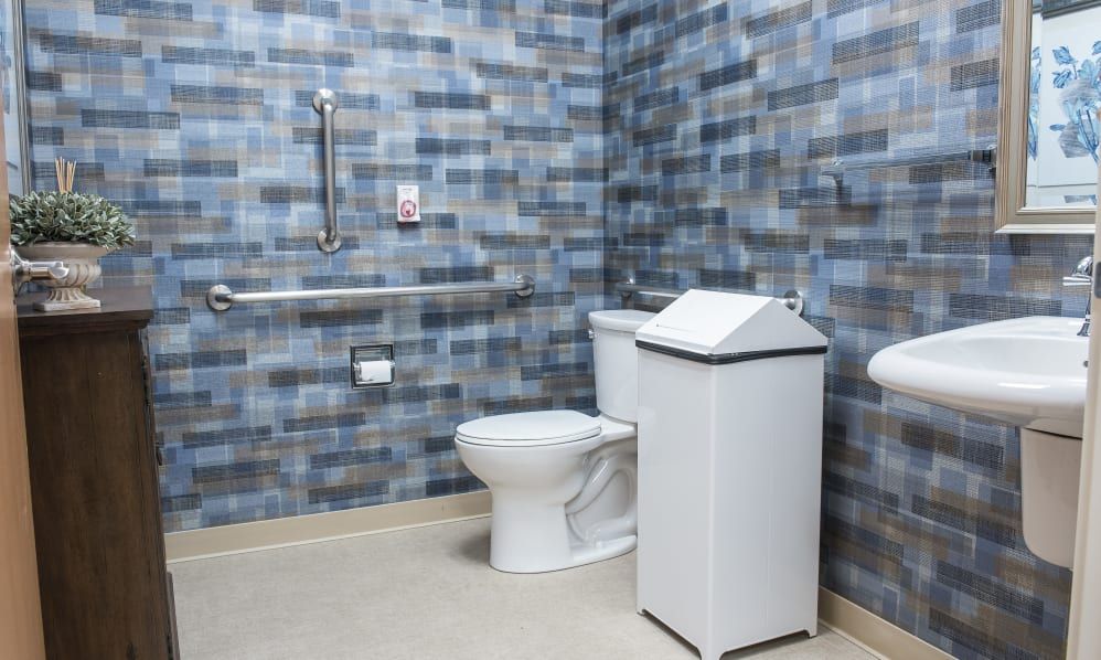 An accessible bathroom with a toilet, grab bars, and a trash can against a tiled wall.