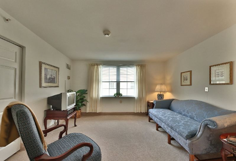 A cozy living room with a sofa, armchair, and television, softly lit by a lamp near the window.