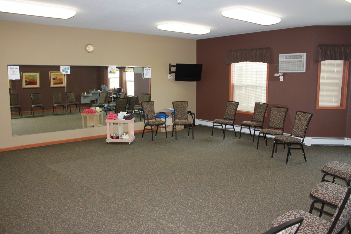 A spacious room with chairs arranged in a semi-circle, featuring mirrors, a wall-mounted television, and windows.