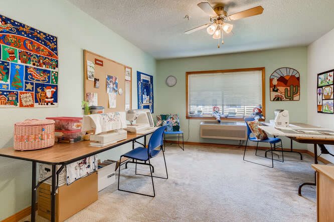 A bright classroom with desks and chairs arranged next to large windows, featuring a bulletin board and a ceiling fan.