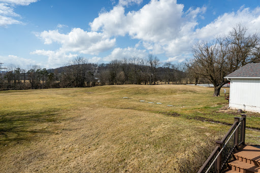A serene view of an open field under a partly cloudy sky, surrounded by trees and distant hills.