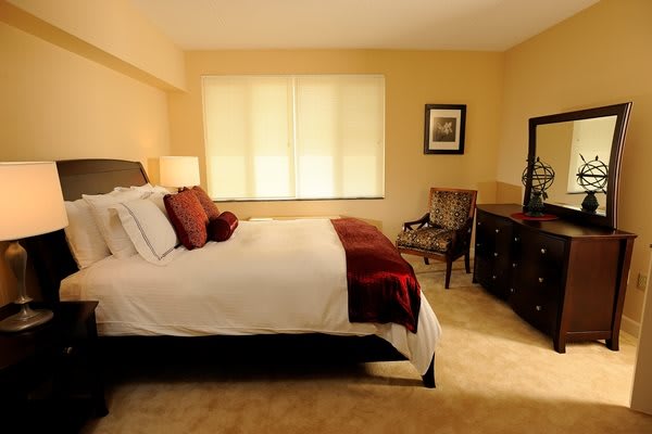 A cozy bedroom featuring a neatly made bed with red and white accents, a window allowing natural light, and classic wooden furniture.