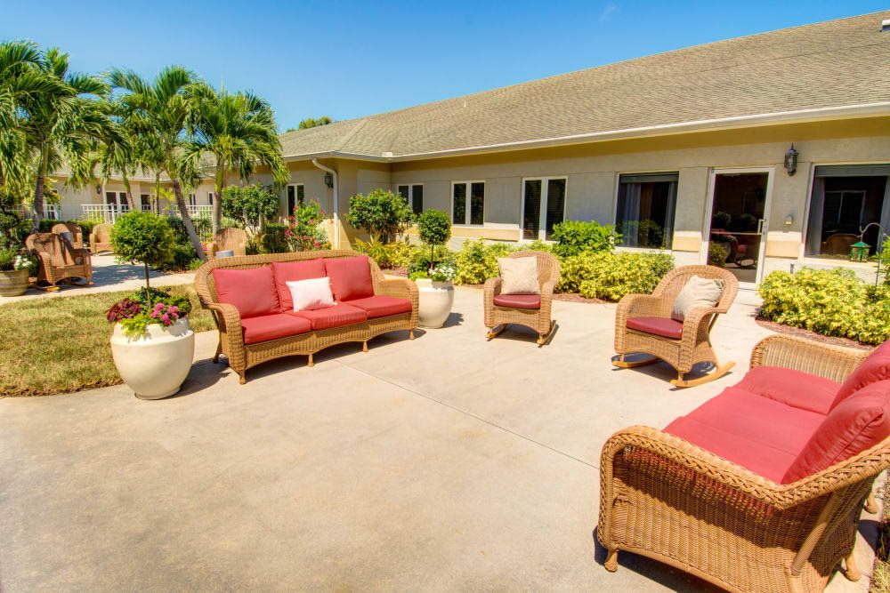 A sunny patio with wicker furniture and red cushions, surrounded by lush greenery.