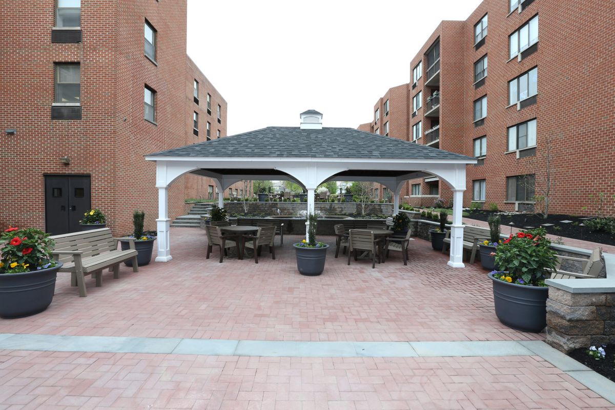 Outdoor gazebo area with tables, benches, and potted plants surrounded by brick buildings.