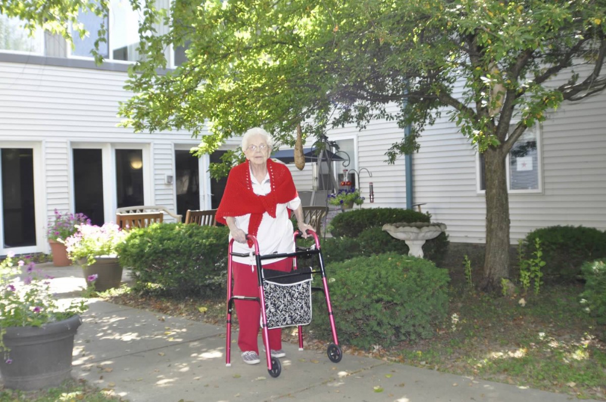 An elderly woman using a walker outside a house with trees in the background.