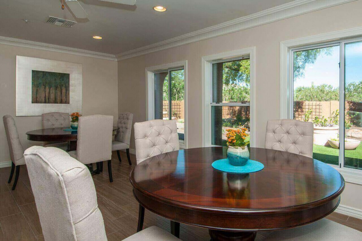 A cozy dining room with a round wooden table and upholstered chairs, surrounded by windows letting in natural light.