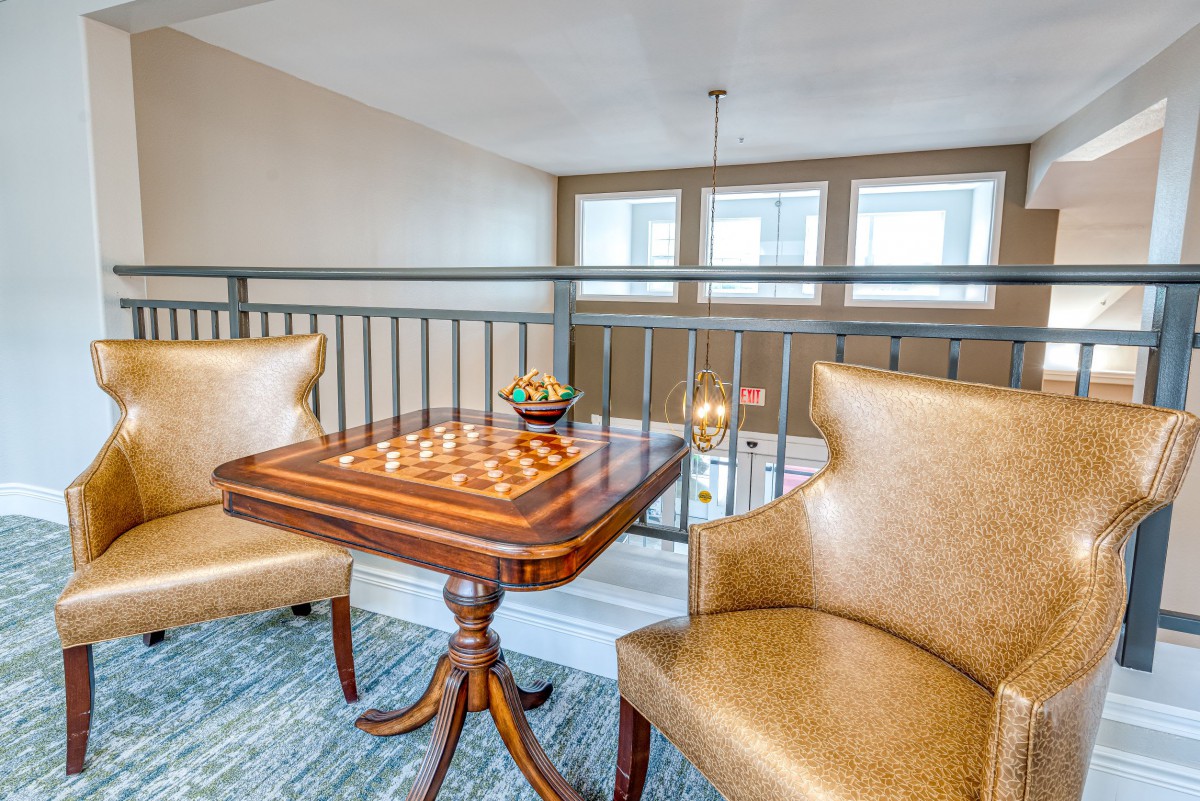A cozy lounge area with a chessboard set on a small table between two chairs, bathed in natural light from the nearby windows.