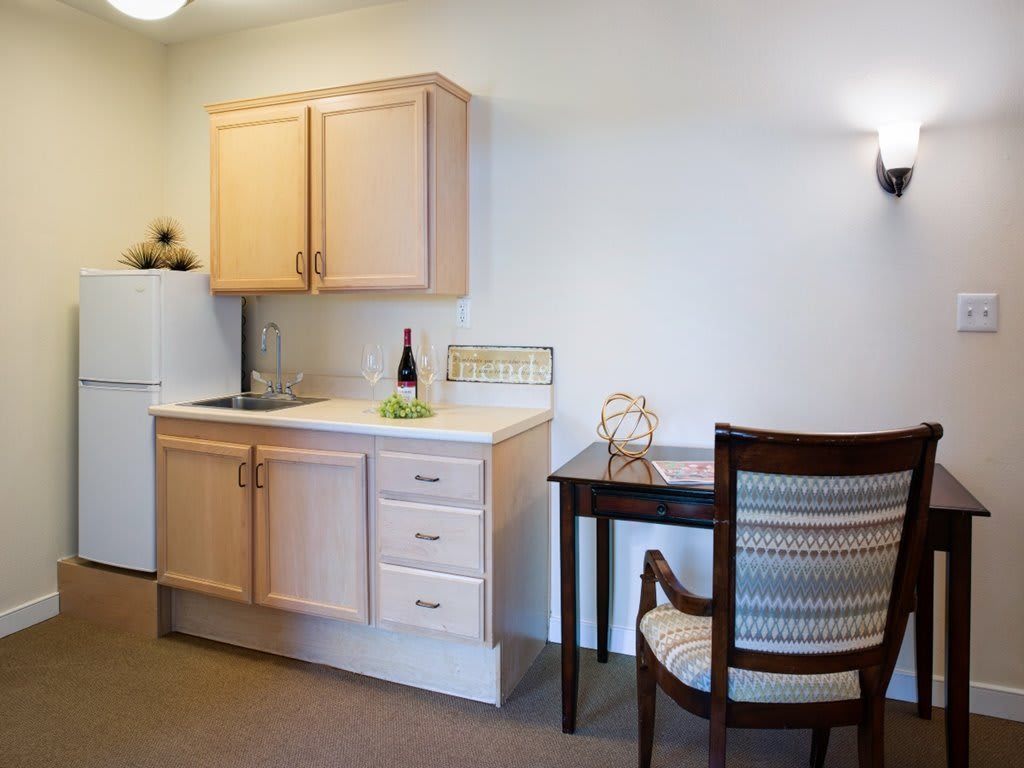 A compact kitchenette with wooden cabinets, a small desk, and a chair in a cozy indoor setting.