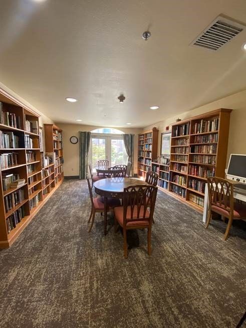 A cozy library reading room with bookshelves lined along the walls, a central table surrounded by chairs, and a computer station.