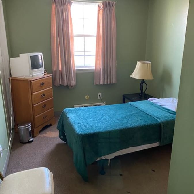 A cozy bedroom with a single bed, nightstand, lamp, and a dresser with a TV.
