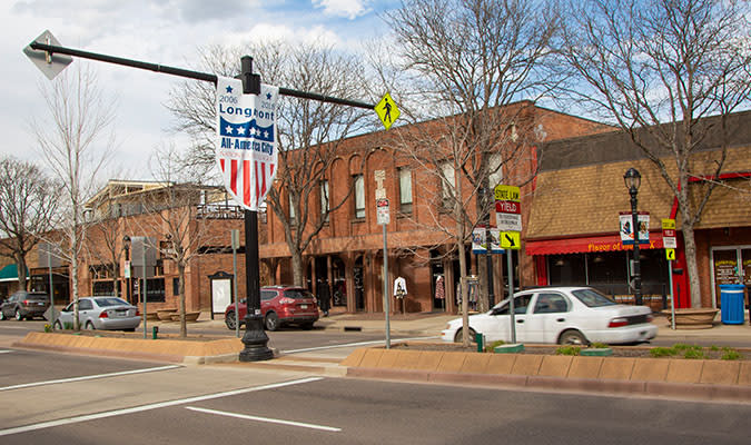 A view of a downtown street with brick buildings, cars parked along the road, and a crosswalk in the foreground.