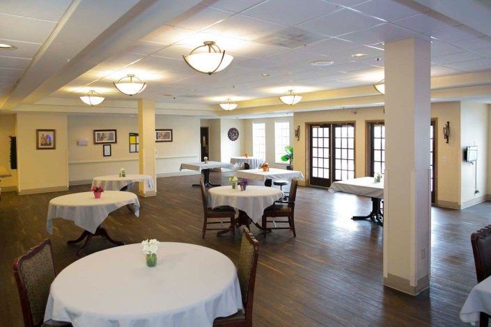 A well-lit dining area with round tables and chairs, covered with white tablecloths.