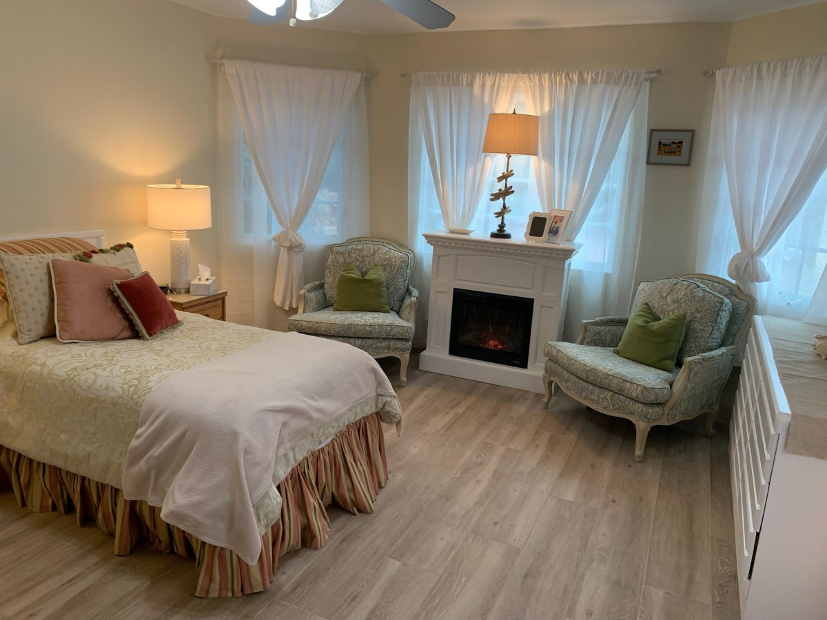 A cozy bedroom featuring a comfortable bed, two armchairs, and a fireplace, all set on a wooden floor with soft lighting.