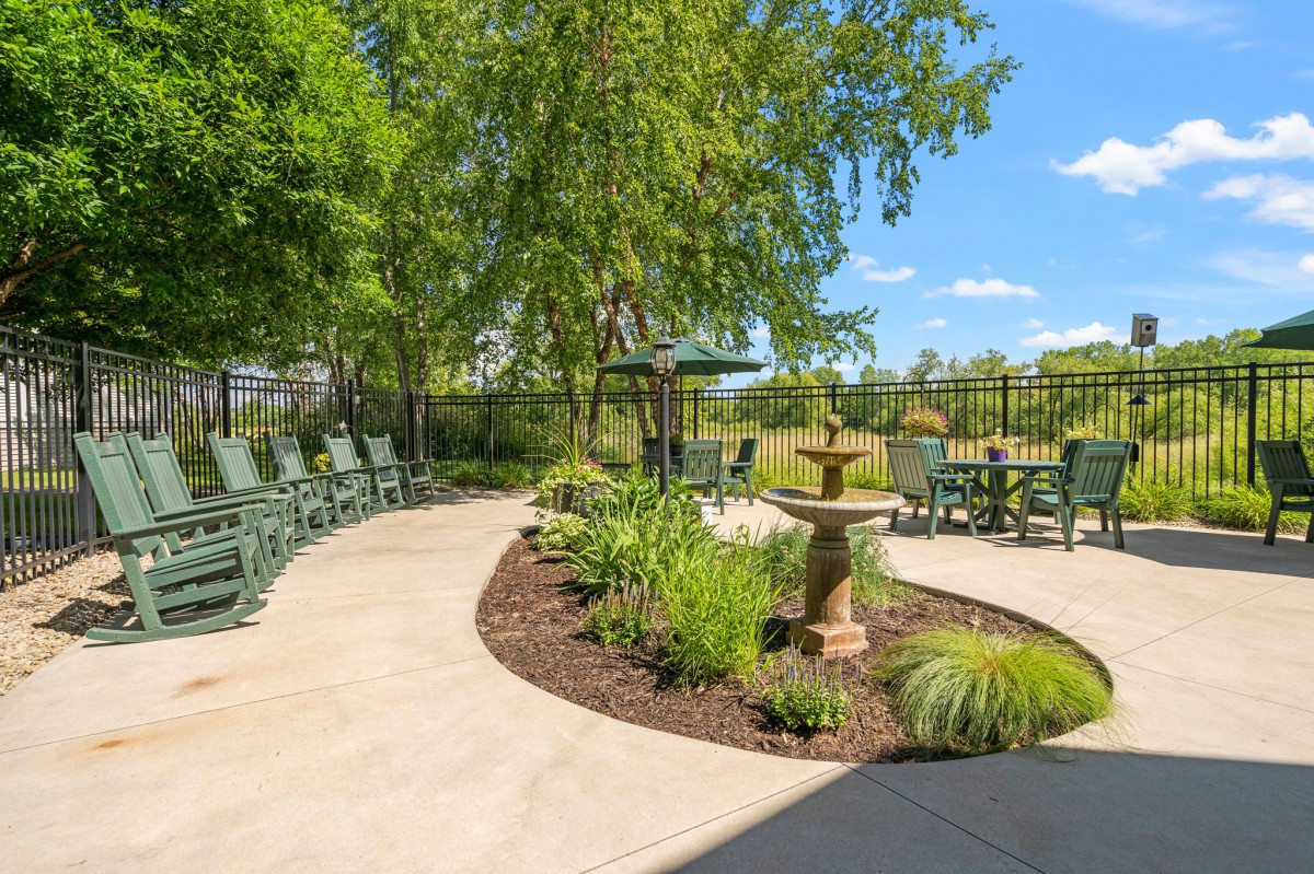 A tranquil outdoor garden with a patio, fountain, and seating under trees on a sunny day.
