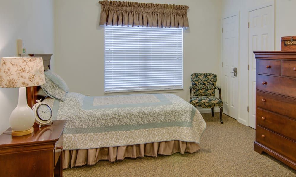 A cozy bedroom with a single bed covered by a quilt, a window with blinds and curtains, and a patterned chair in the corner.