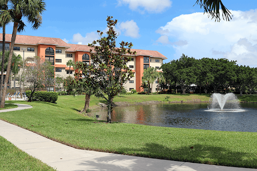 A serene view of an apartment complex by a tranquil pond, surrounded by lush greenery and a clear blue sky.