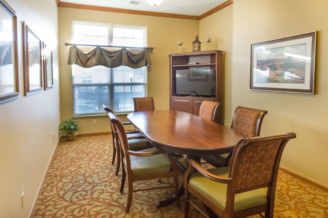 A conference room with a long wooden meeting table surrounded by chairs, a television on a cabinet, and large windows with curtains.