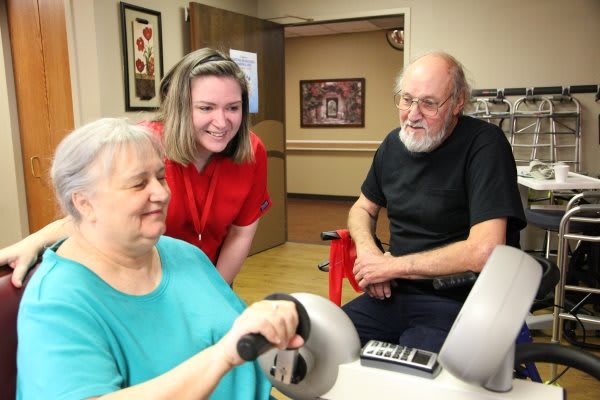 An elderly woman participates in a therapy session with the assistance of a healthcare worker, while another person observes.