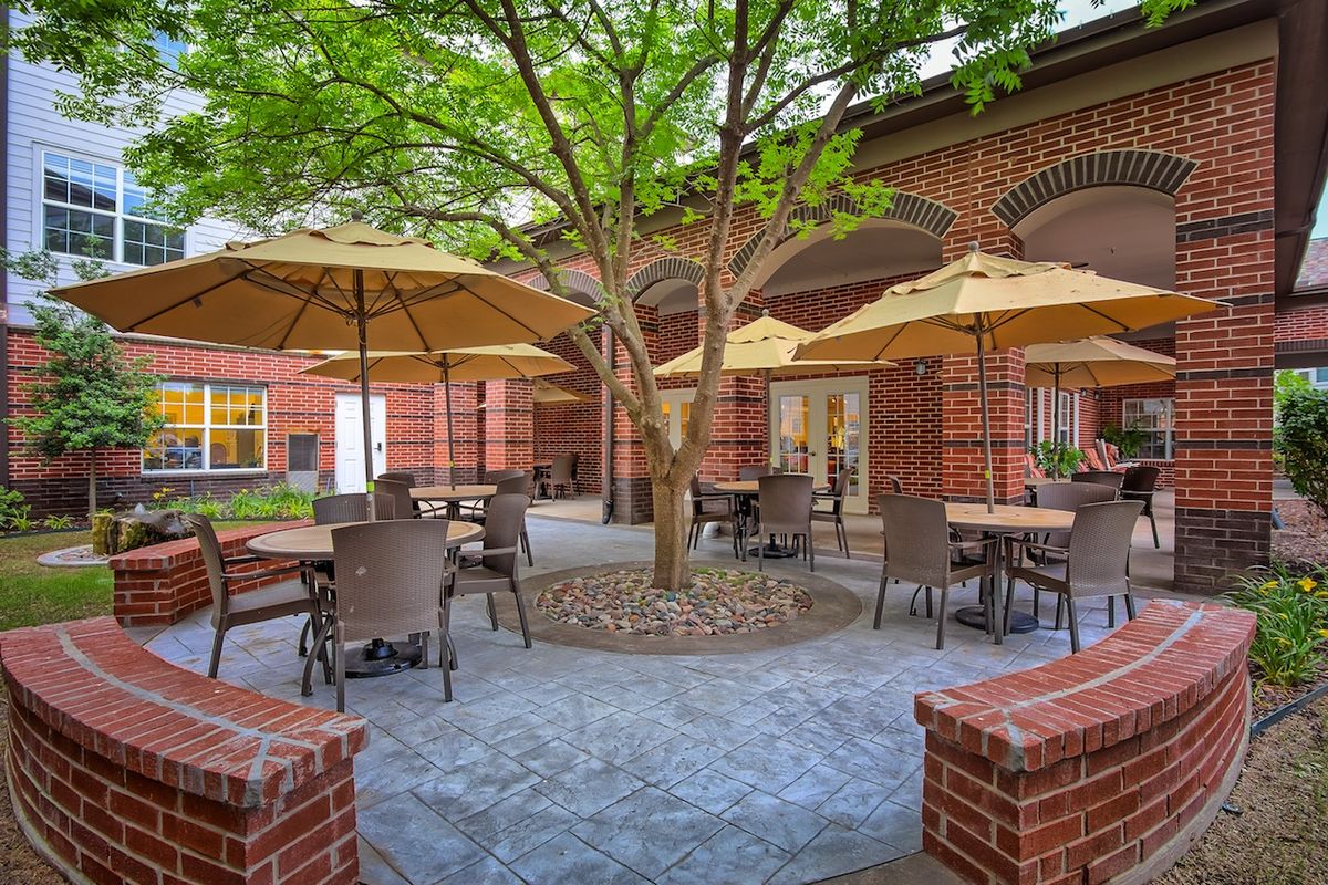 A tranquil courtyard with outdoor seating, featuring tables and umbrellas under a tree surrounded by brick architecture.