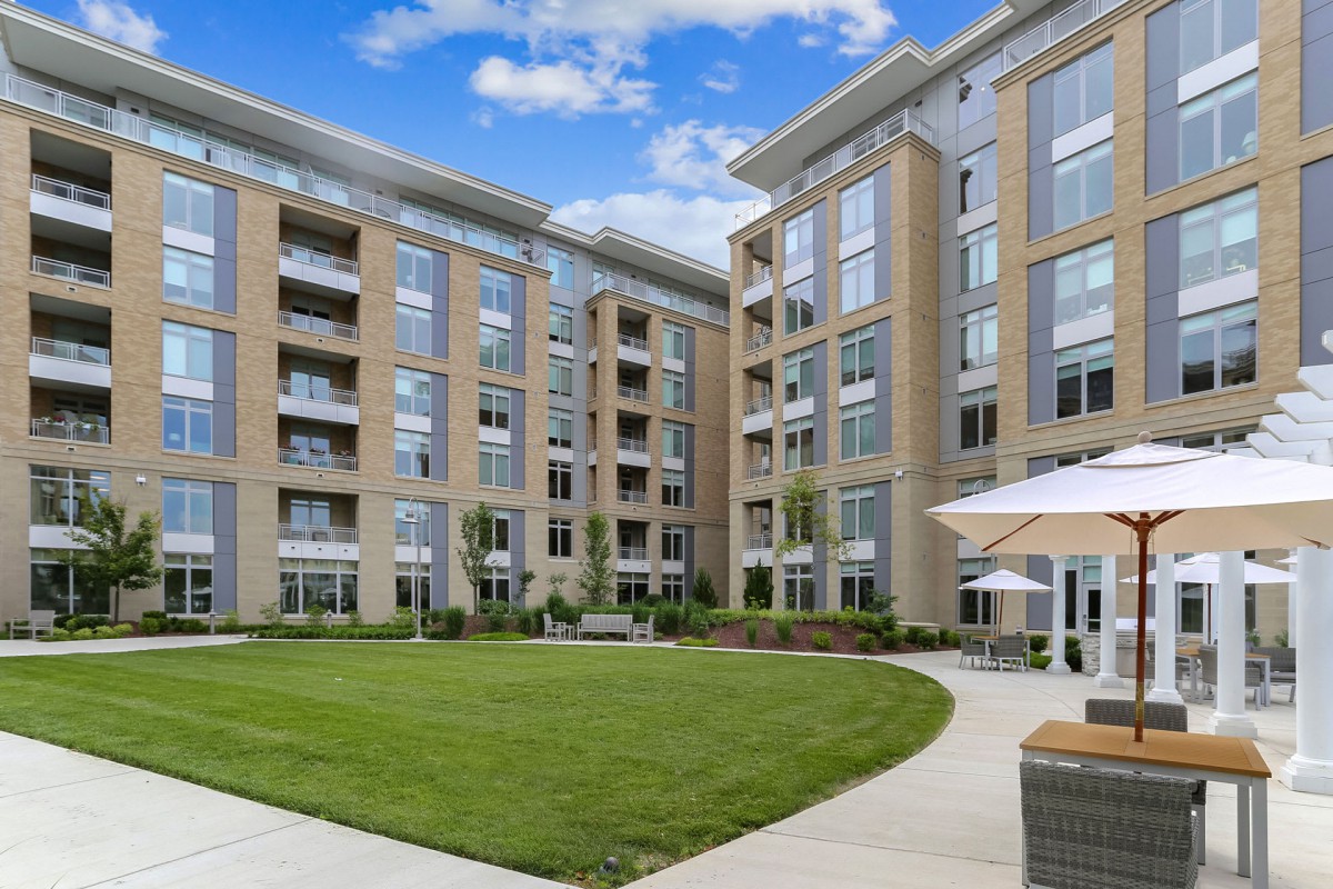 A modern apartment building with a well-maintained courtyard and green lawn, featuring outdoor seating and umbrellas.