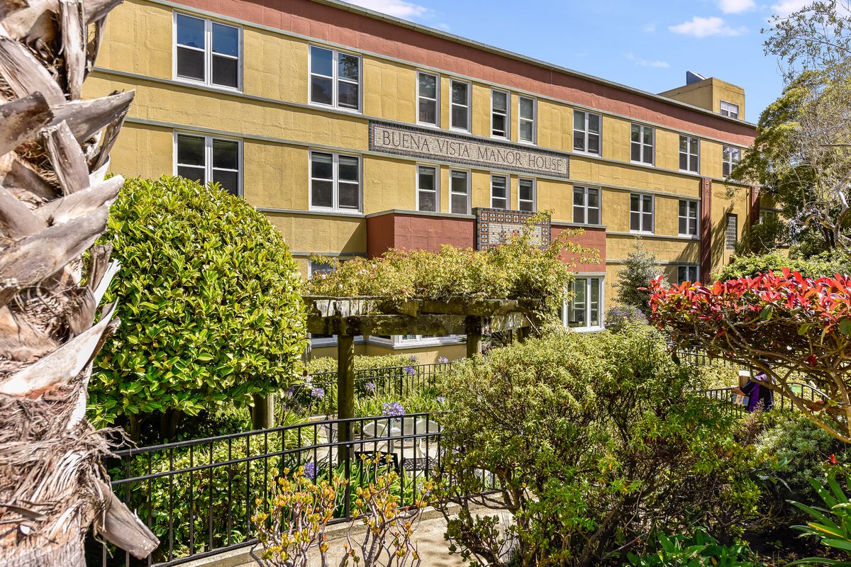 A hospital building with a garden and trees in the foreground.